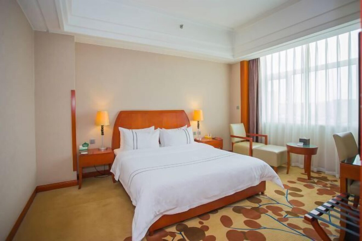 A hotel room with a large bed, two bedside lamps, a chair, a small table, and a window with curtains.