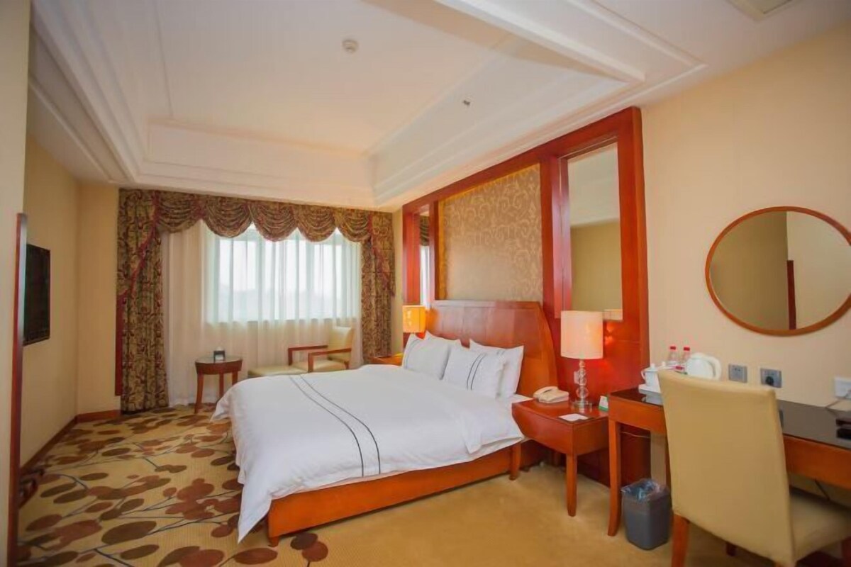 A hotel room with a large bed, a desk with a chair, a round mirror, and a small table.