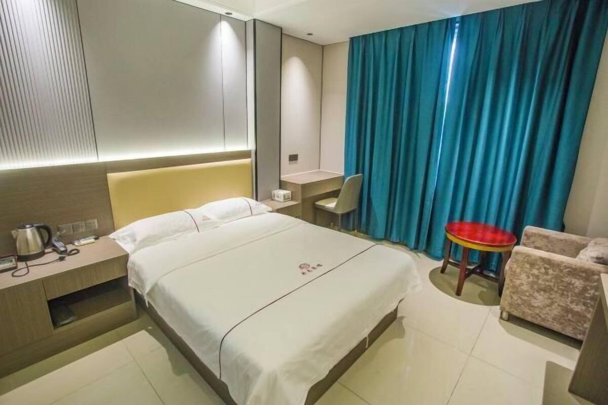 A hotel room with a bed, bedside table with a kettle, a desk, a chair, a small table, and a blue curtain.