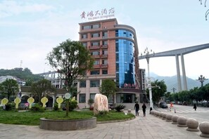 Un hotel de varios pisos con fachada curva, zona de estar al aire libre con mesas redondas y un árbol grande en el centro.