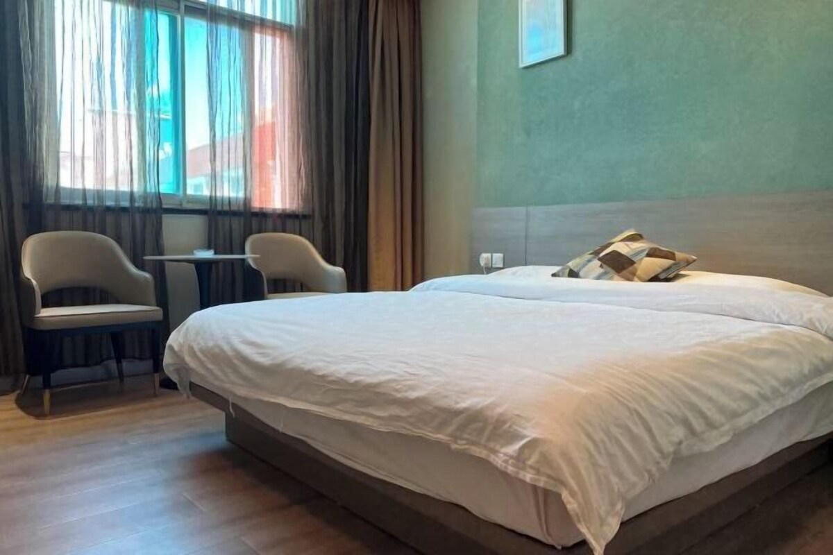 A hotel room with a large bed, two chairs, a small table, and a view of the outside through a window with curtains.