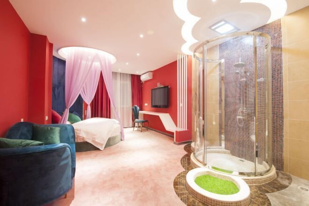 A hotel room with a bed, a blue armchair, a desk, a TV, and a glass-enclosed shower area.