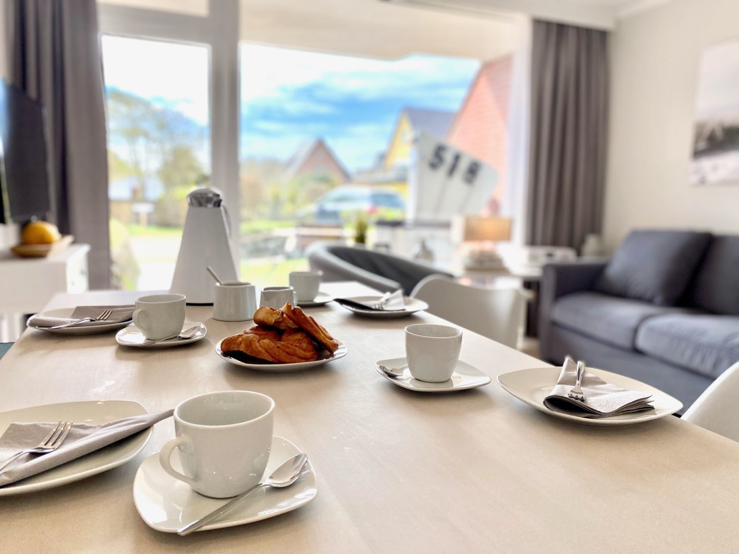 Ein Esstisch ist gedeckt mit Tellern, Tassen und Besteck in einem modernen Zimmer mit einer Couch und Blick auf Häuser.