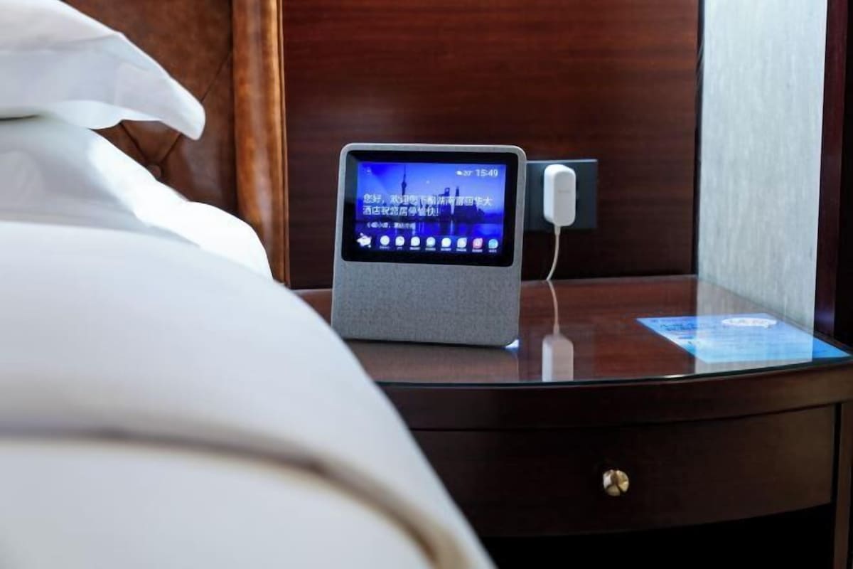 Une chambre d’hôtel avec une table de chevet sur laquelle se trouve une horloge numérique et un petit écran, un tiroir de table de chevet et un lit avec du linge de lit blanc.