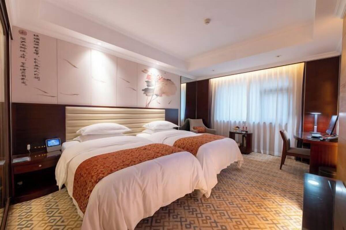 A hotel room with two beds, a desk, a chair, a TV, and a large window with curtains.