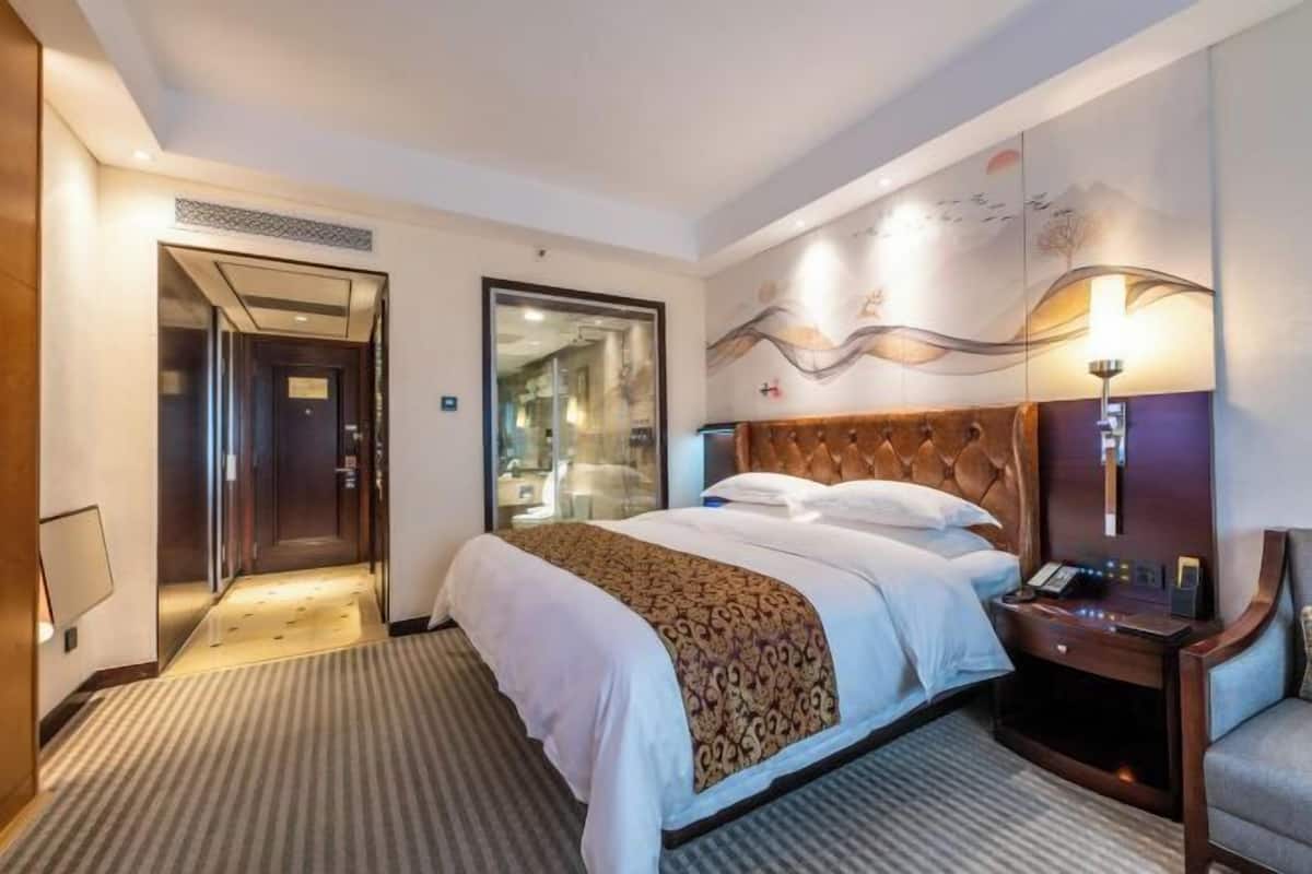 A hotel room with a large bed, a wooden headboard, a bedside table with a lamp, and a view of the hallway.