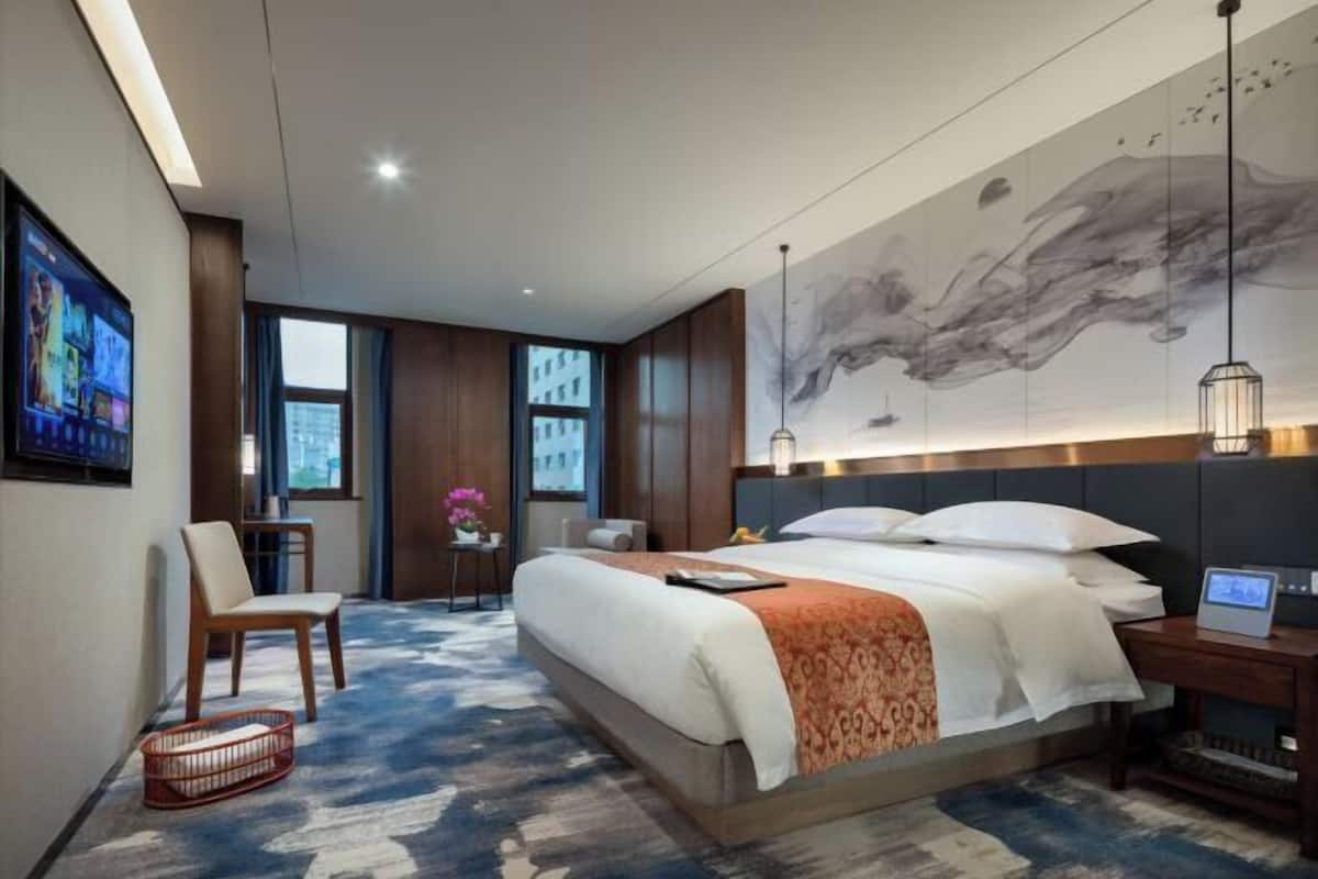 A modern hotel room with a large bed, a desk, a chair, and a large window.