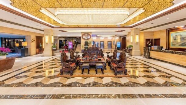 Een ruime lobby met een sierlijke centrale tafel en stoelen, een receptiebalie en een hoog plafond met decoratieve patronen.