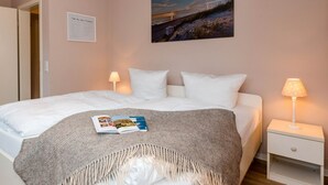 Un lit bien fait, avec un magazine, deux lampes de chevet, un tableau encadré au mur et une petite table de chevet blanche.