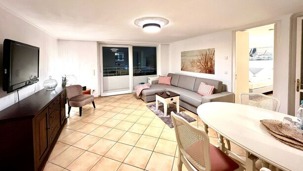 A modern living room with a flat-screen TV, a sofa, a dining table, and a balcony with a view. - Penthouse-Berta - Modern vacation apartment in Westerland: Perfect for your vacation. (Sylt)