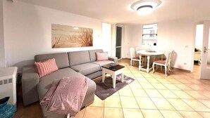 A modern living room with a grey sofa, a small round table, and a dining area with white chairs. - Penthouse-Berta - Modern vacation apartment in Westerland: Perfect for your vacation. (Sylt)