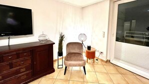 A living room with a flat-screen TV, a wooden cabinet, a beige armchair, a small table with a lamp, and a sliding glass door. - Penthouse-Berta - Modern vacation apartment in Westerland: Perfect for your vacation. (Sylt)
