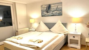 A neatly made bed with pillows, a painting of sailboats on the wall, and two bedside lamps. - Penthouse-Berta - Modern vacation apartment in Westerland: Perfect for your vacation. (Sylt)