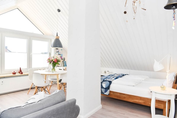 A modern, well-lit room with a skylight, a bed with a blue quilt, a small table with a lamp, and a chair.