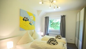 A hotel room with a bed, a bedside table with a lamp, a window with curtains, and a wall with a colorful painting. - Brise-Sylt - The vacation apartment "Brise-Sylt" is situated at ground level and quietly in (Wenningstedt-Braderup (Sylt))