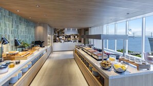 A modern breakfast buffet with a variety of food items, a kitchen area in the background, and a view of the water through large windows. - Hilton Garden Inn Yokohama Minatomirai (Yokohama)
