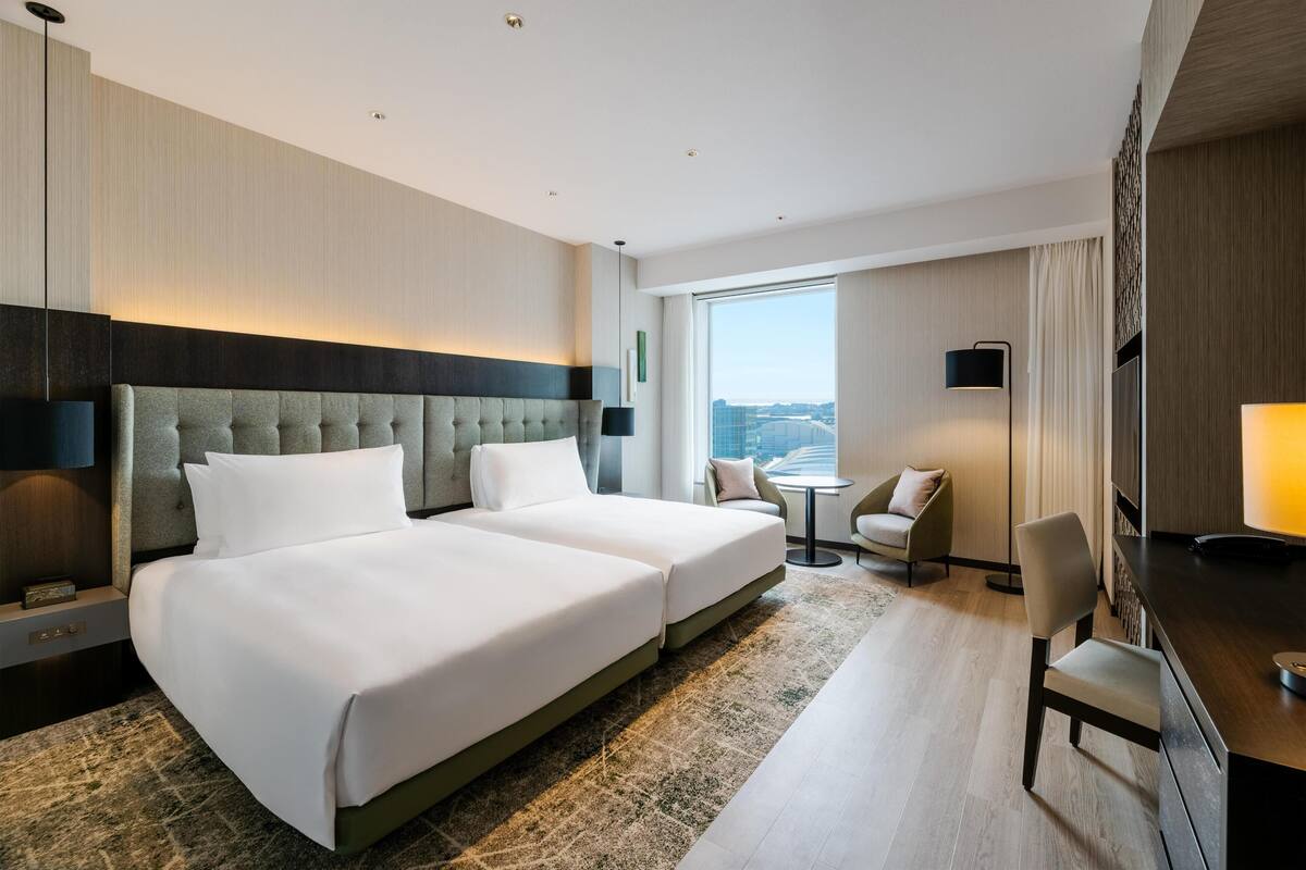 Ein Hotelzimmer mit zwei Betten, einem Schreibtisch, einem Stuhl, einem kleinen Tisch und einem großen Fenster mit Blick auf die Stadt.