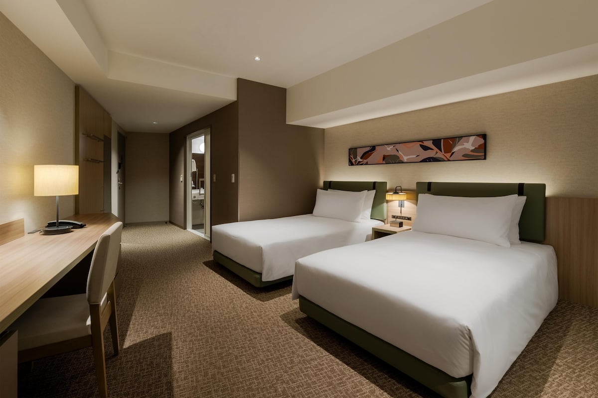 Ein Hotelzimmer mit zwei Betten, einem Schreibtisch, einem Sessel, einer Lampe und einem Bild an der Wand.