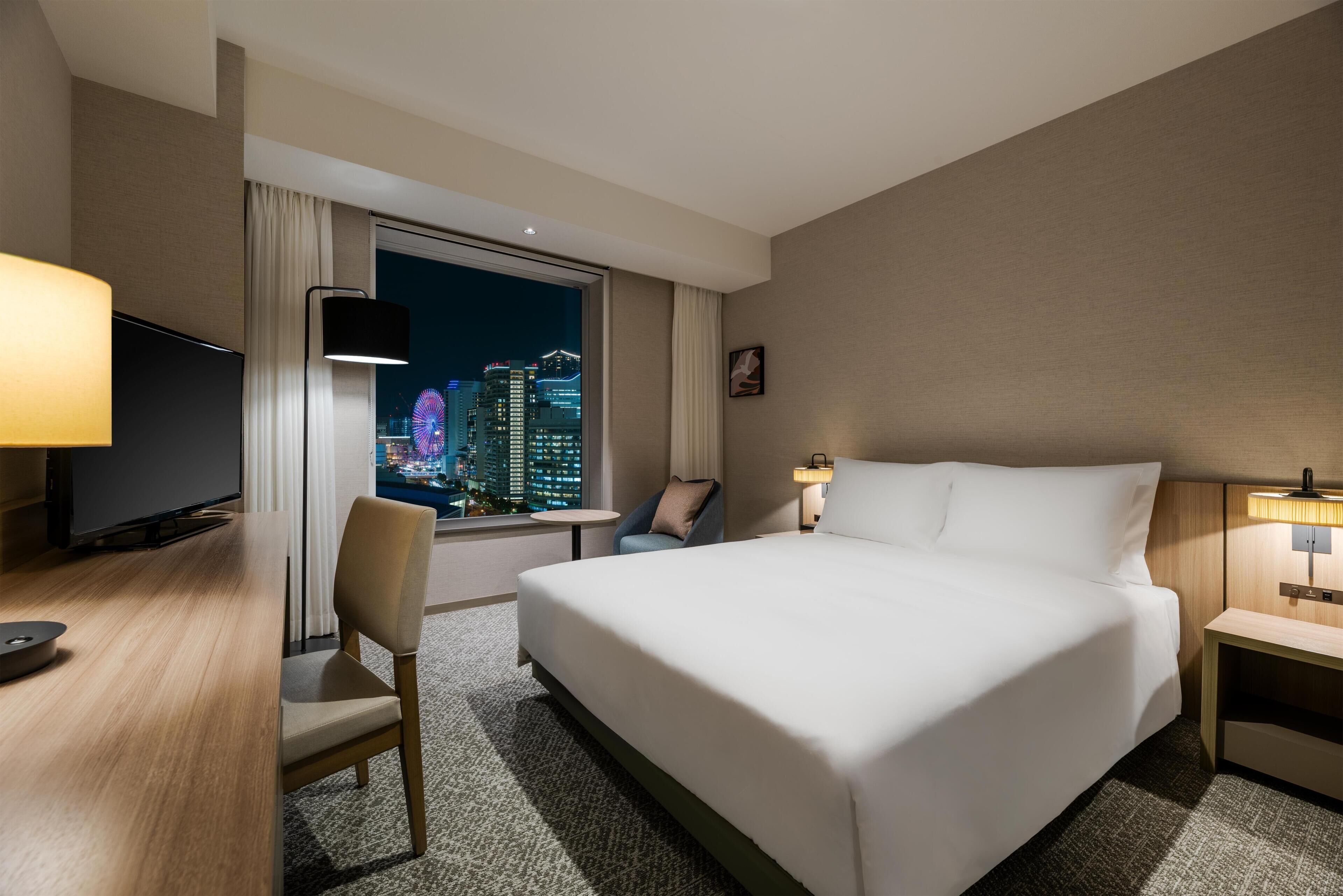 A hotel room with a bed, a desk, a chair, a TV, a window with a city view, and a nightstand.