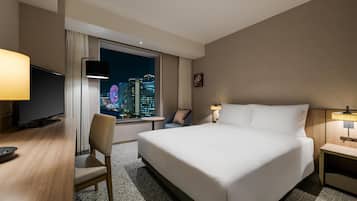 A hotel room with a bed, a desk, a chair, a TV, a window with a city view, and a nightstand.