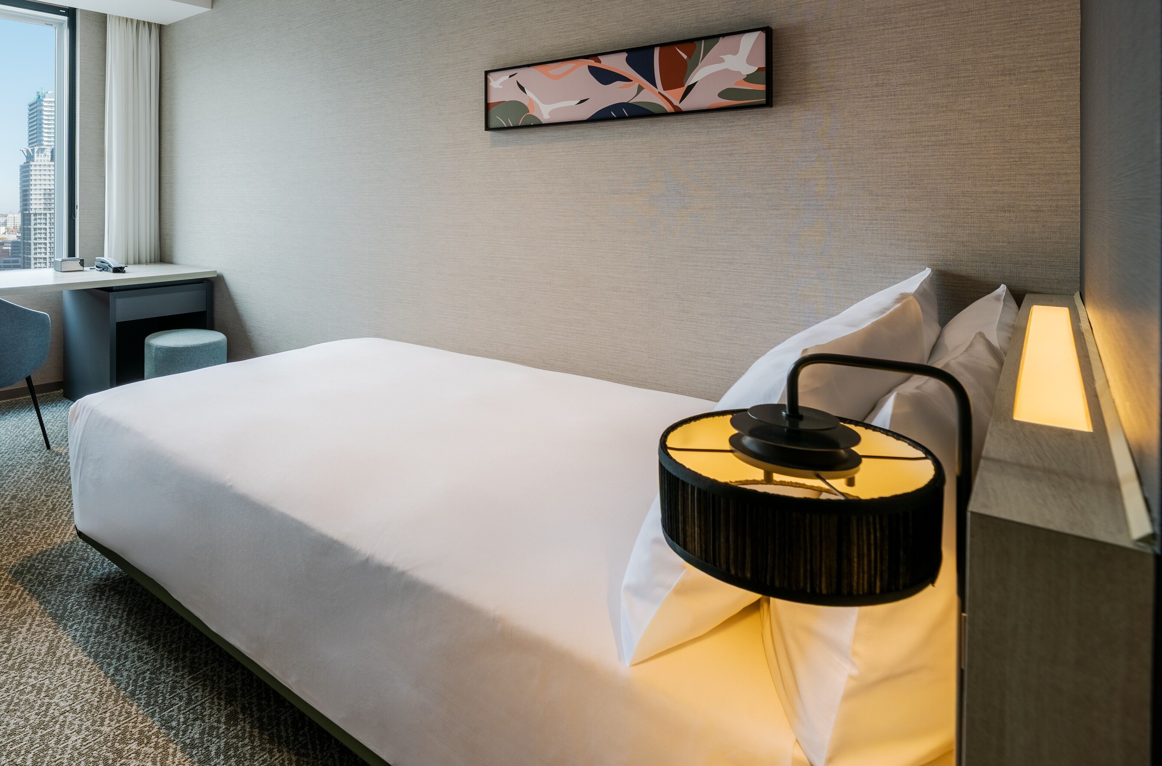 Quarto de hotel com cama, escrivaninha, cadeira, abajur e vista para a cidade.