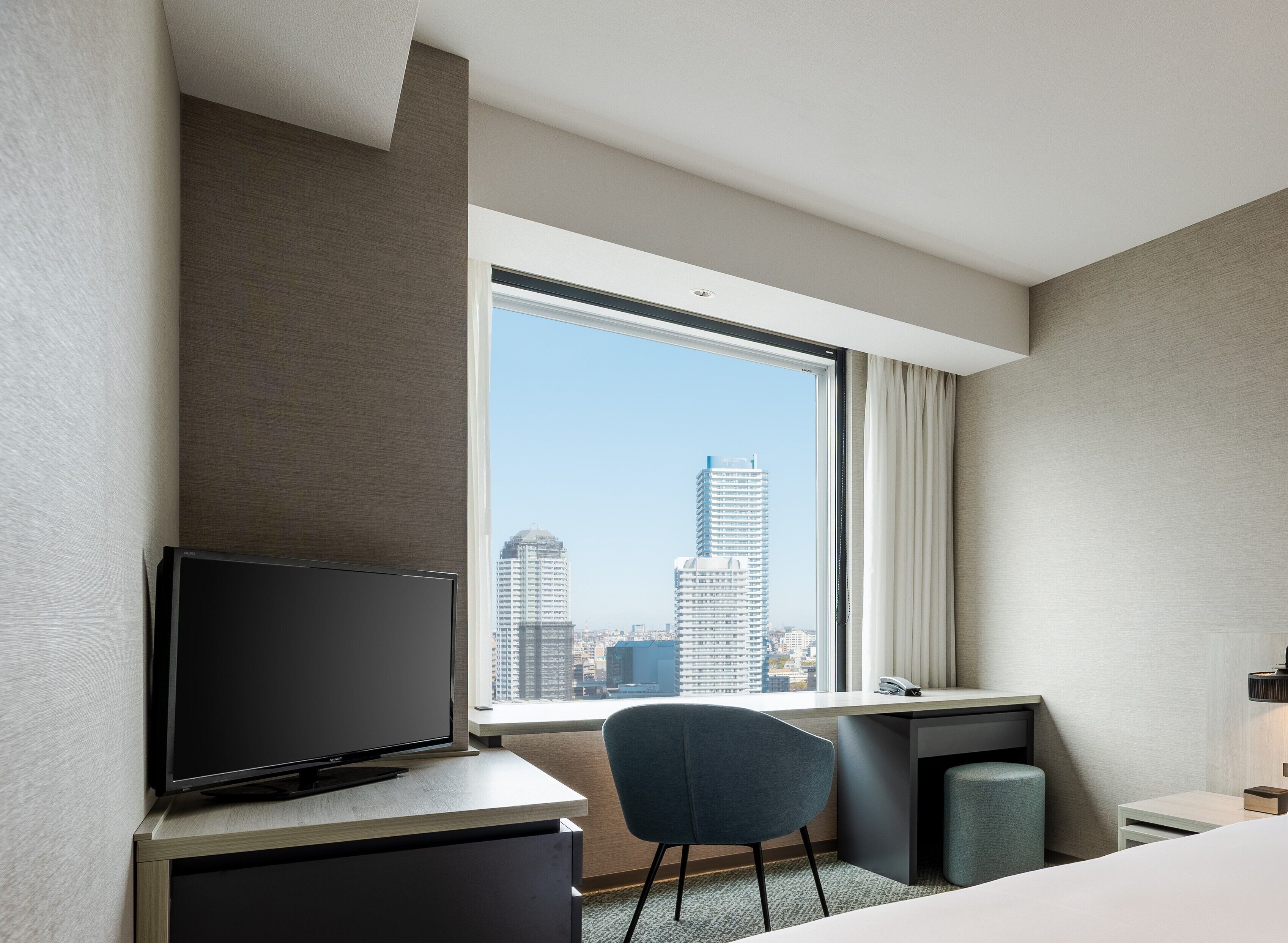 A hotel room with a desk, chair, television, and a large window with a city view.