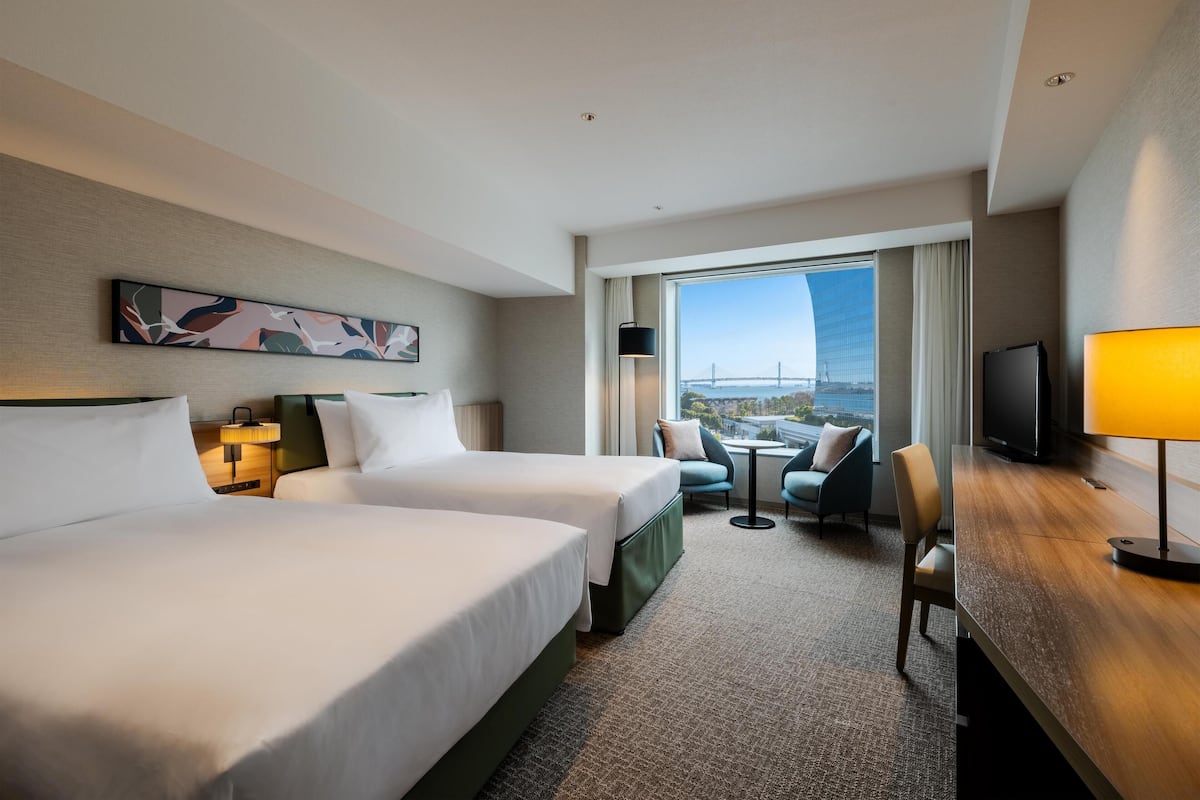 Ein Hotelzimmer mit zwei Betten, einem Schreibtisch, einem Stuhl, einem Fernseher und Blick auf eine Brücke.