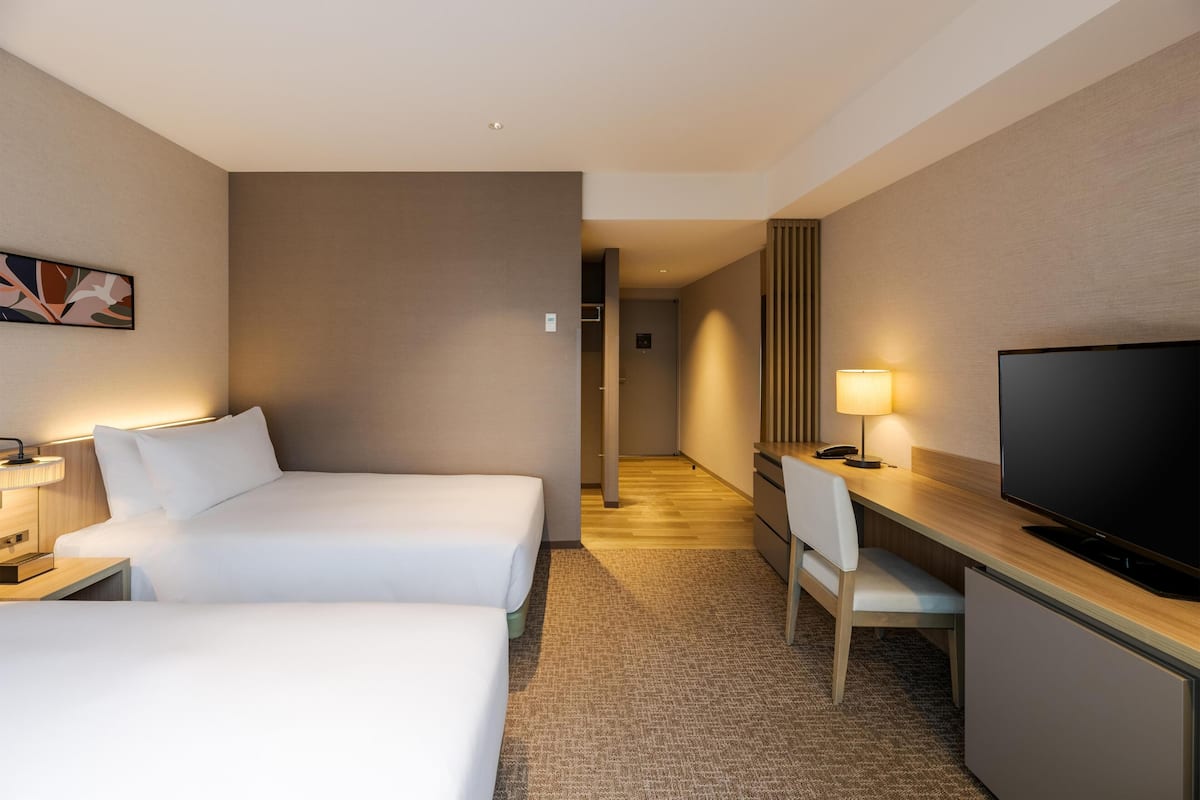 Ein Hotelzimmer mit zwei Betten, einem Schreibtisch mit Fernseher, einem Sessel und einer Lampe.