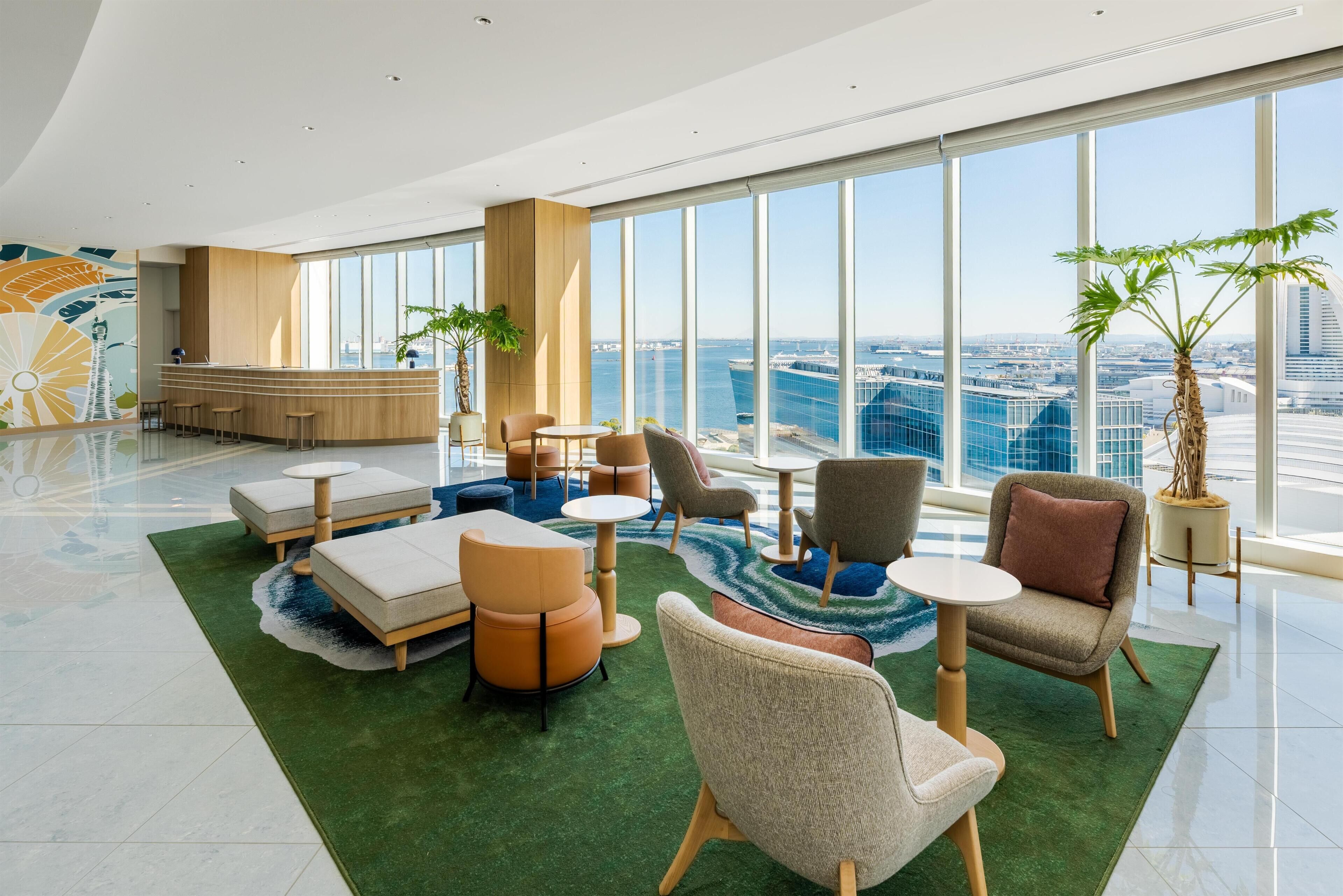 A modern hotel lobby with a reception desk, seating area, large windows, and a view of the cityscape.