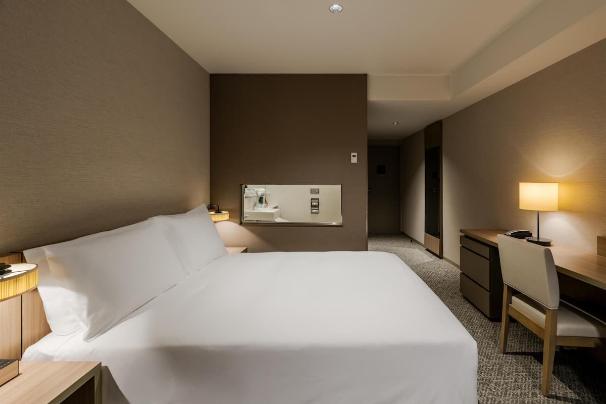 Ein Hotelzimmer mit Bett, Nachttisch, Lampe, Schreibtisch, Stuhl und Spiegel.