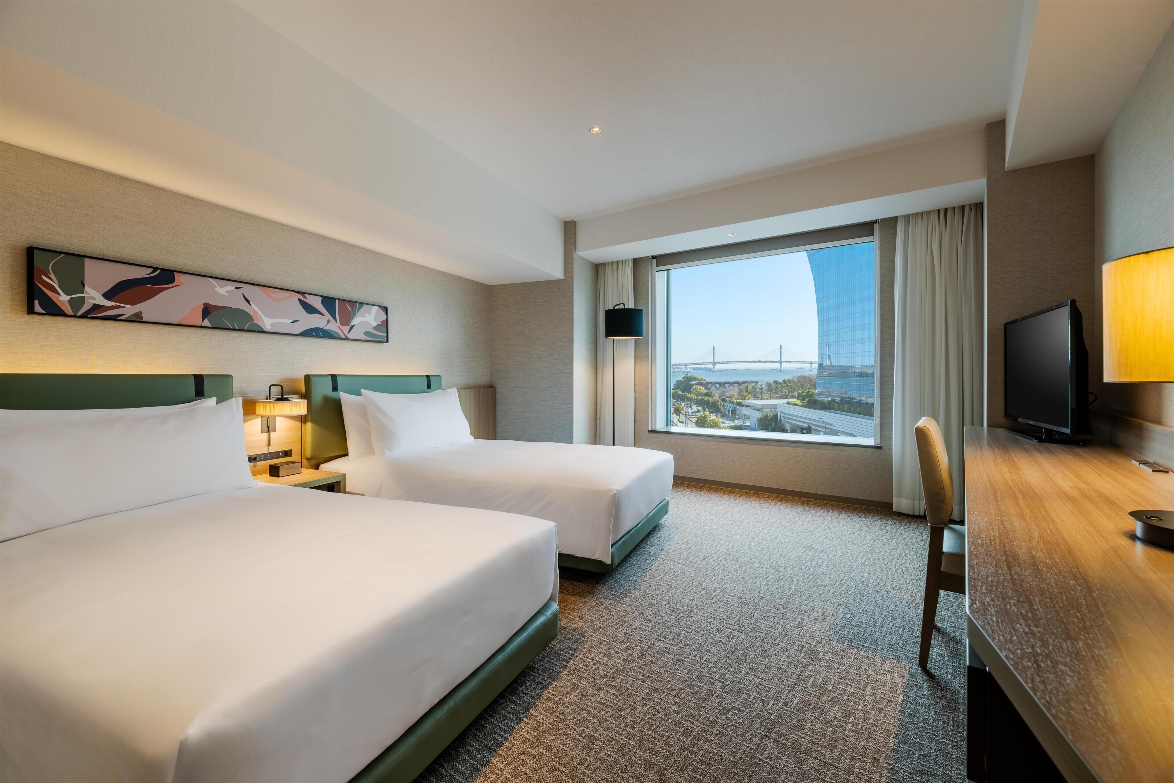 A hotel room with two beds, a desk, a TV, a large window with a view, and a painting on the wall.