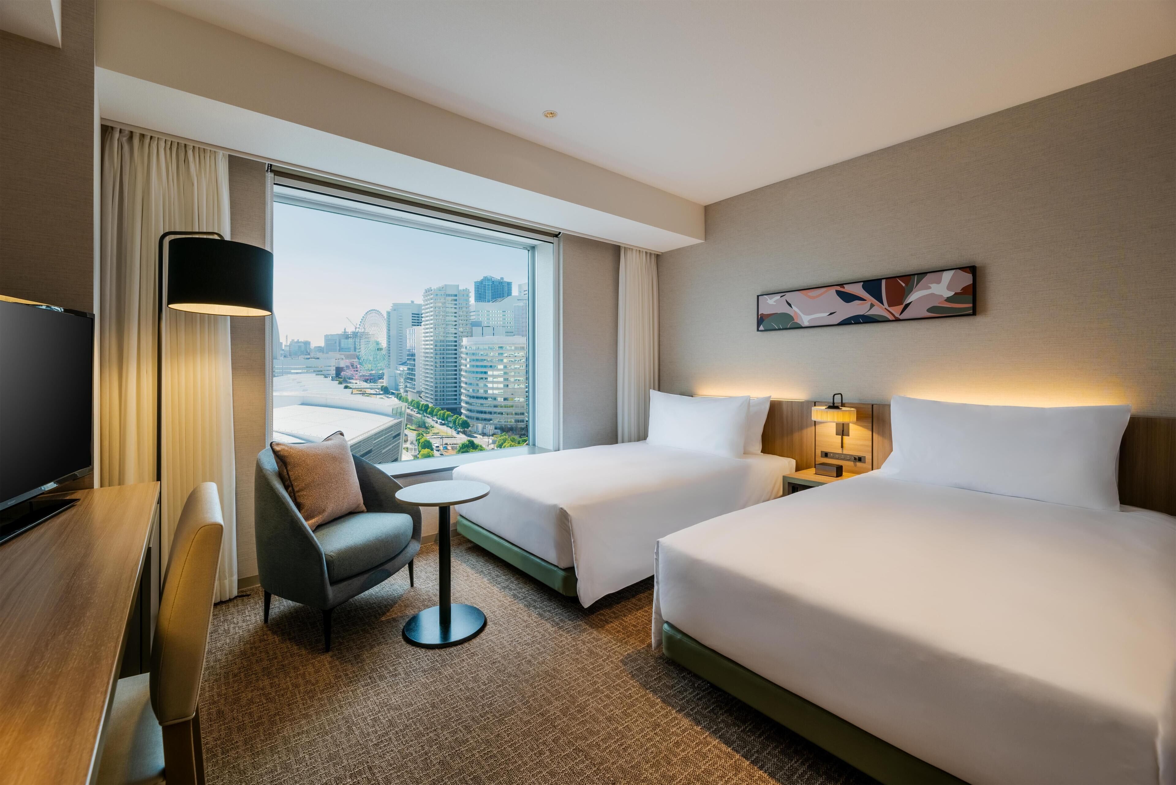 Quarto de hotel com duas camas, uma escrivaninha, uma cadeira, uma mesinha e uma televisão.