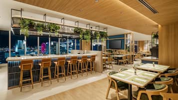 Zona de bar moderna con taburetes de madera, una barra y plantas colgantes.