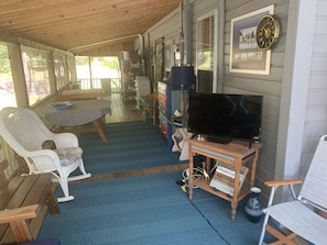 Une véranda abritée avec une table en bois, des chaises en osier, un téléviseur posé sur un meuble en bois et un tableau encadré accroché au mur.