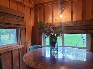 Un intérieur en bois avec une table ronde, un vase rempli de fleurs et une vue sur un paysage verdoyant, visible à travers de grandes fenêtres.