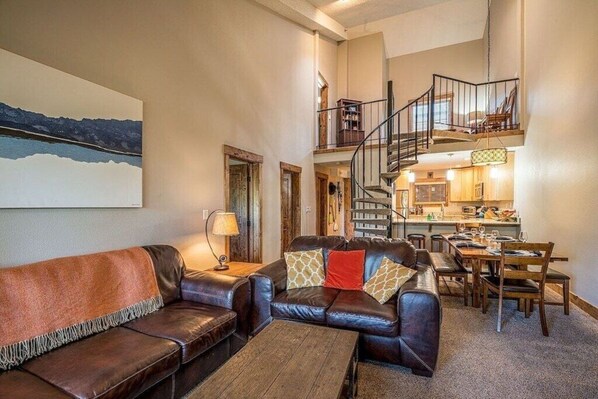 A spacious living area with a leather sofa, a coffee table, and a dining table set for four. A staircase leads to an upper level with a kitchen and dining area.