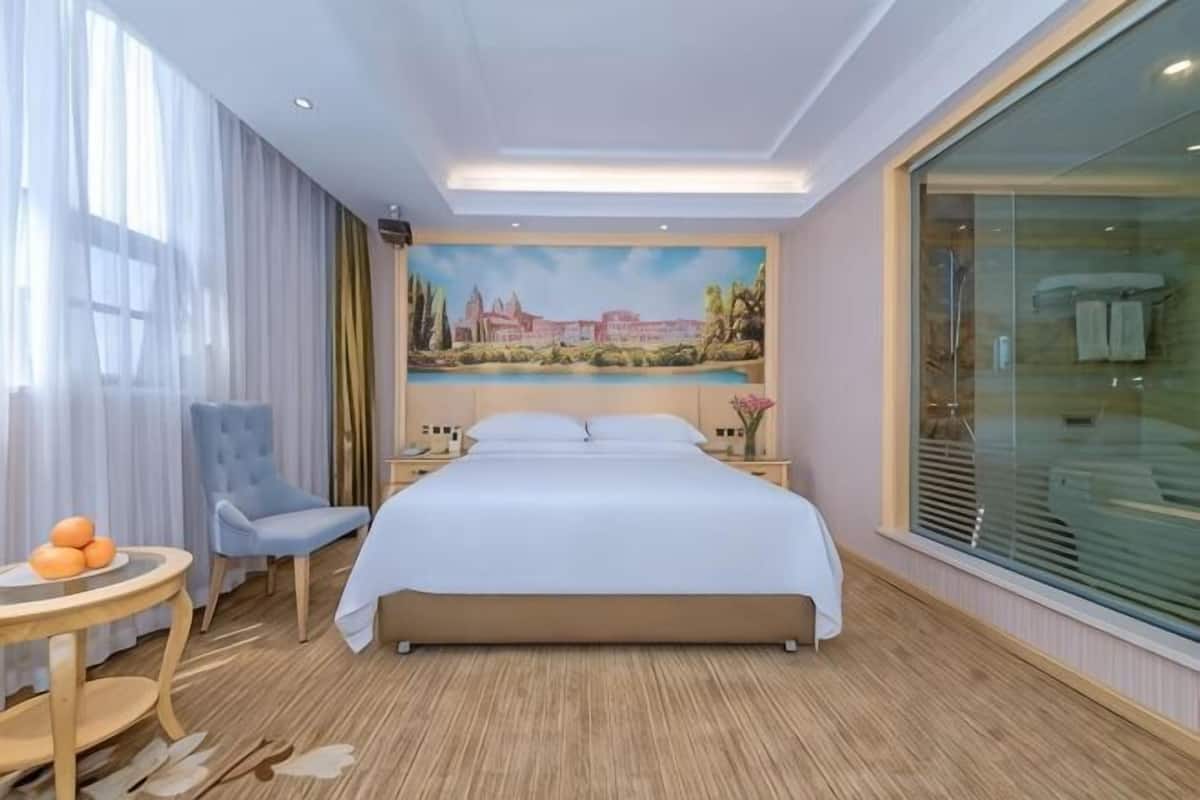 A hotel room with a large bed, a round table with fruit, a chair, and a large window with a view.