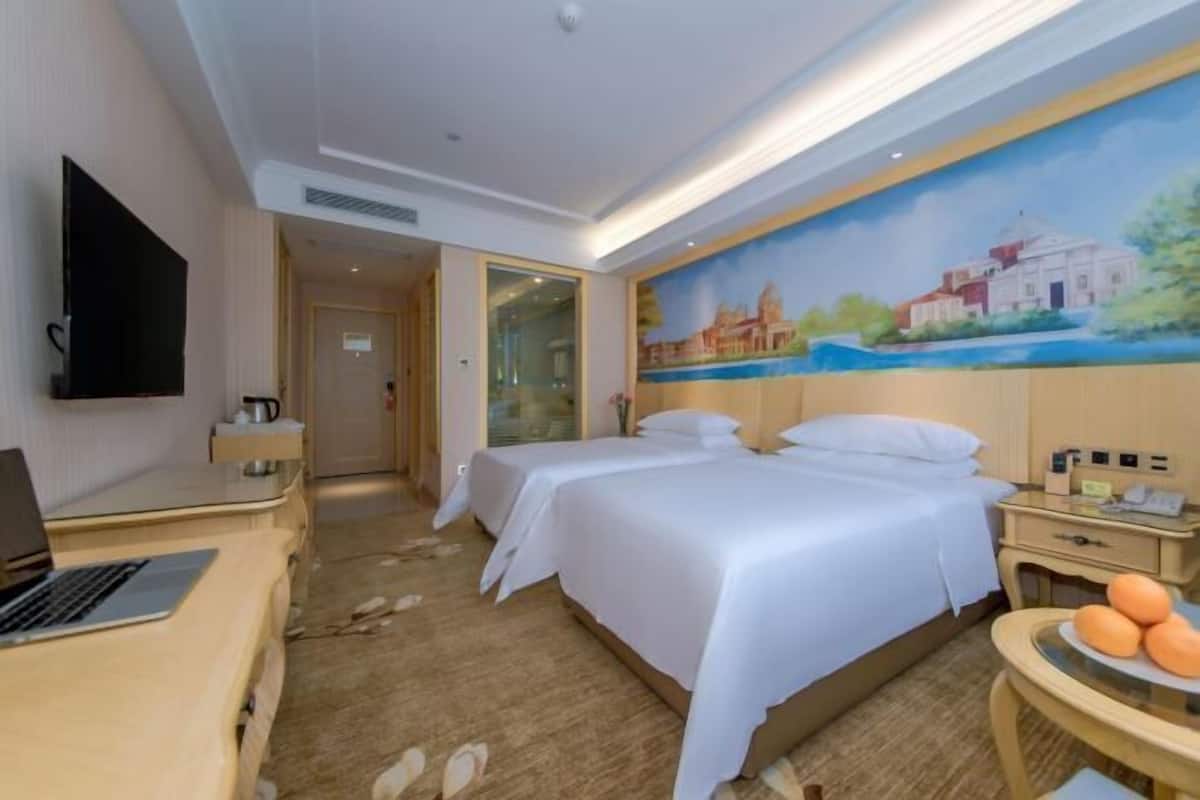 A hotel room with two beds, a desk with a laptop, a TV, and a painting on the wall.