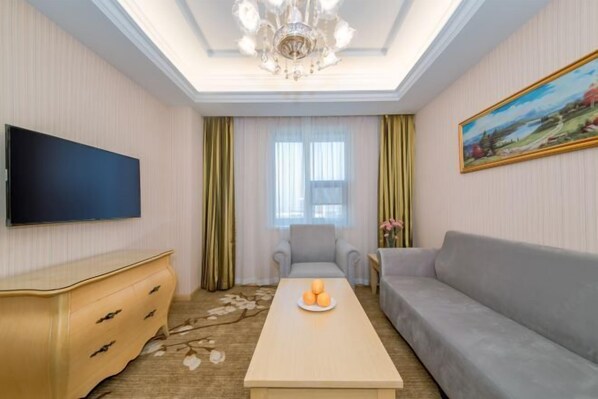 A living room with a flat-screen TV, a sofa, a coffee table with fruit, a chandelier, and a painting on the wall. - Vienna Hotel (Wuhan Sports Center) (Wuhan)