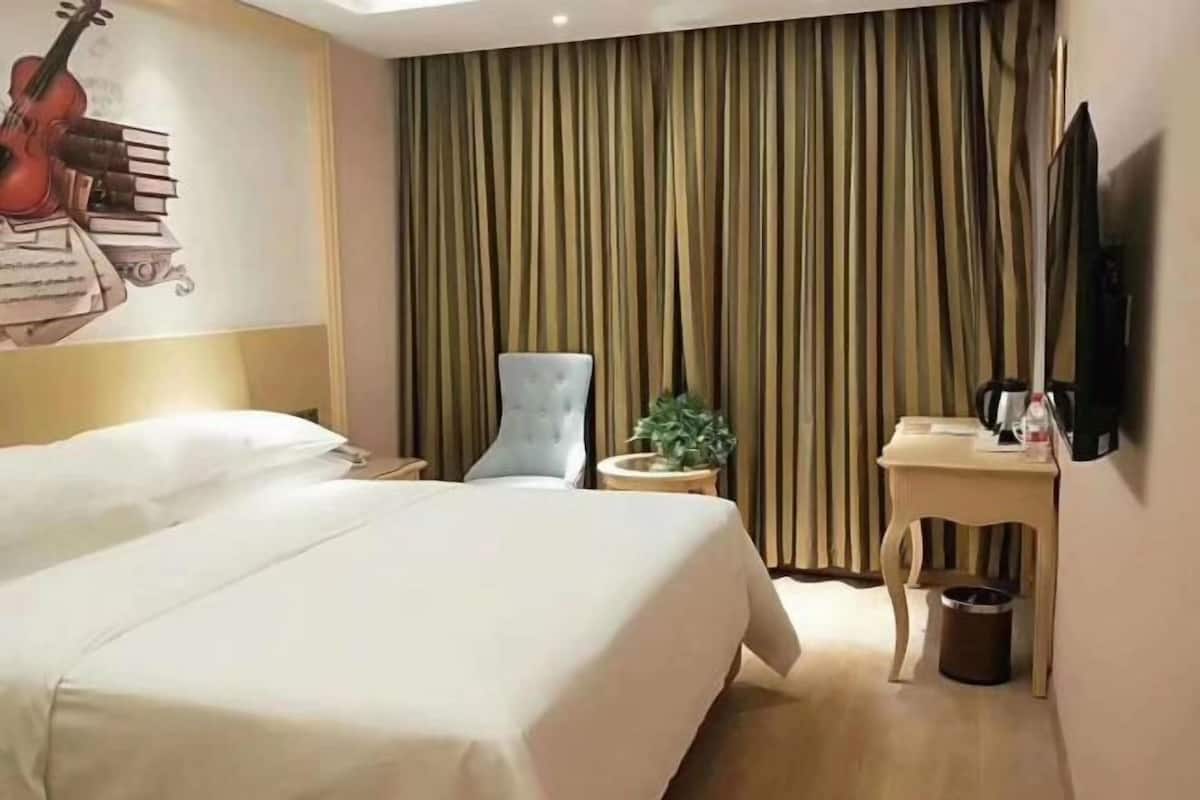 A hotel room with a bed, a chair, a small table, a potted plant, a trash bin, a wall-mounted TV, and a wall decoration of a guitar and books.