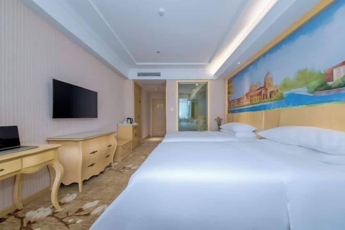 A hotel room with a bed, a desk with a laptop, a TV, and a painting on the wall.