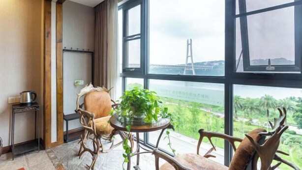 A room with a large window, a wooden chair, a small table with a plant, and a view of a bridge and greenery outside. - Aibao Hotel (Chongqing)