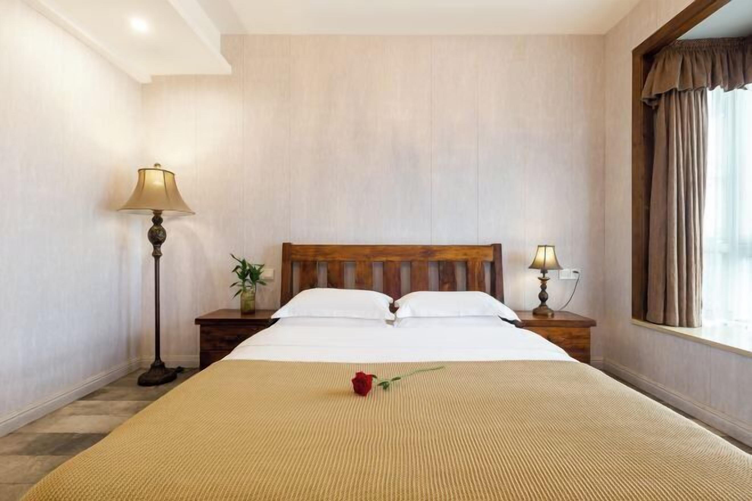 Una habitación de hotel con una cama de madera, dos lámparas de noche, una ventana con cortinas y una sola rosa sobre la cama.