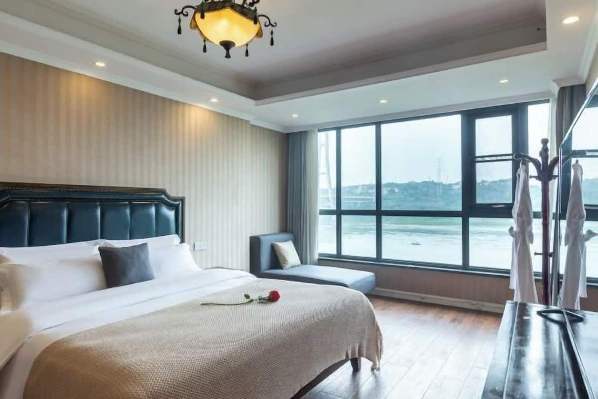 Quarto de hotel com uma cama grande, área de estar, vista para a água e um candelabro.