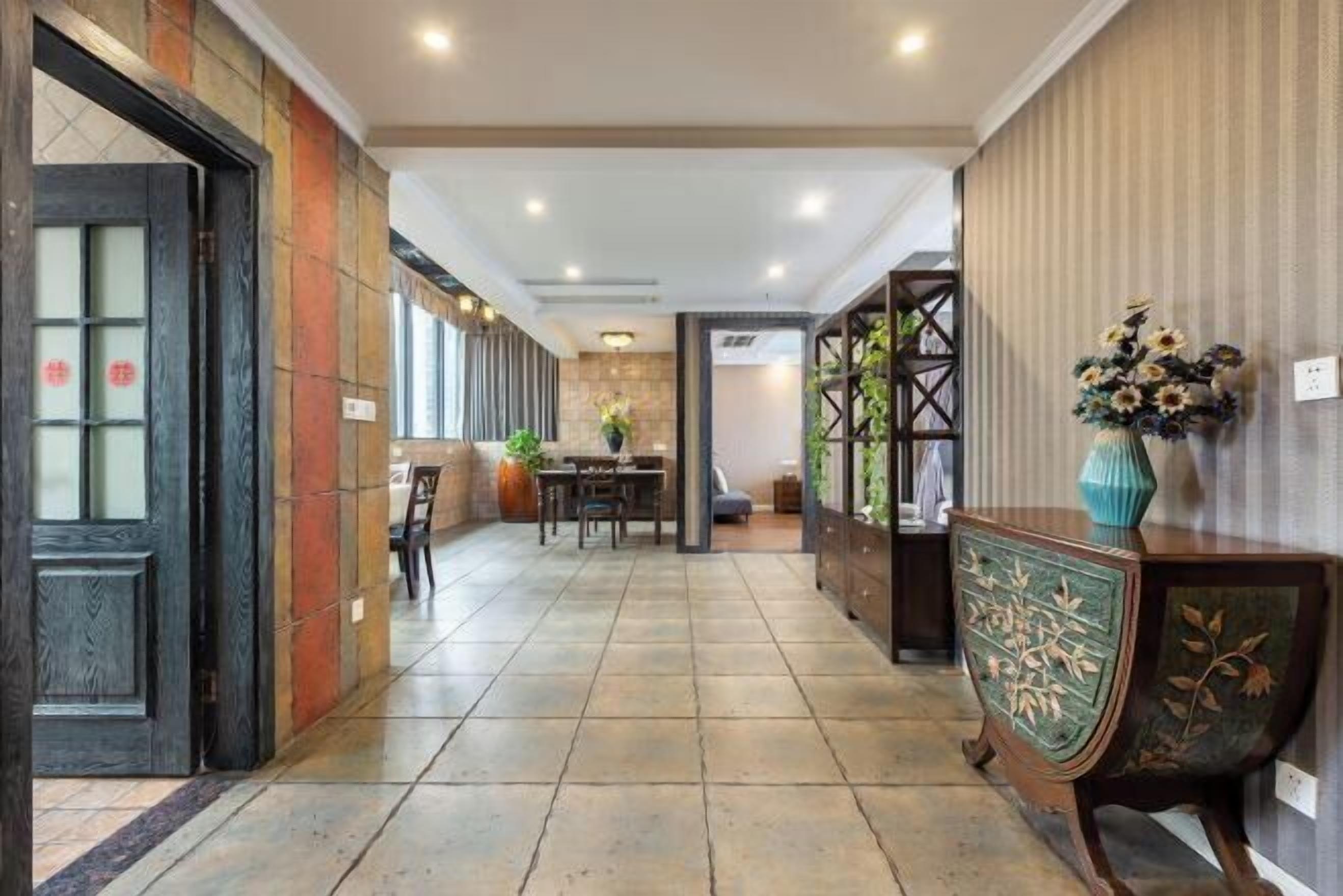 Un pasillo de hotel con suelo de baldosas, una zona de comedor con sillas y una mesa, y un aparador decorativo con un jarrón de flores.