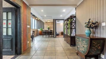 Un pasillo de hotel con suelo de baldosas, una zona de comedor con sillas y una mesa, y un aparador decorativo con un jarrón de flores.