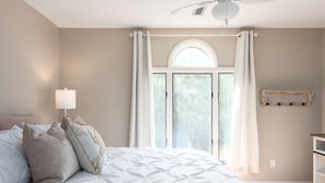 A bedroom with a large bed, a ceiling fan, a window with curtains, and a wall-mounted shelf.