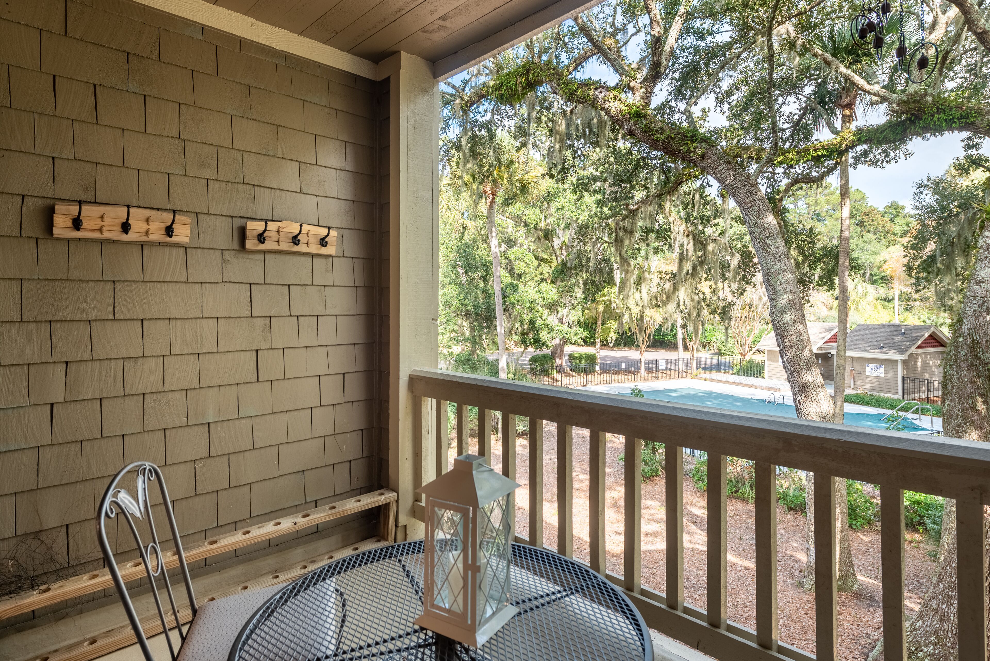 A covered patio with a round table and chairs, a lantern, and a view of a swimming pool and trees.
