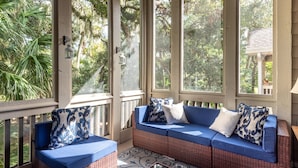 A covered porch with wicker furniture, large windows, and outdoor views.
