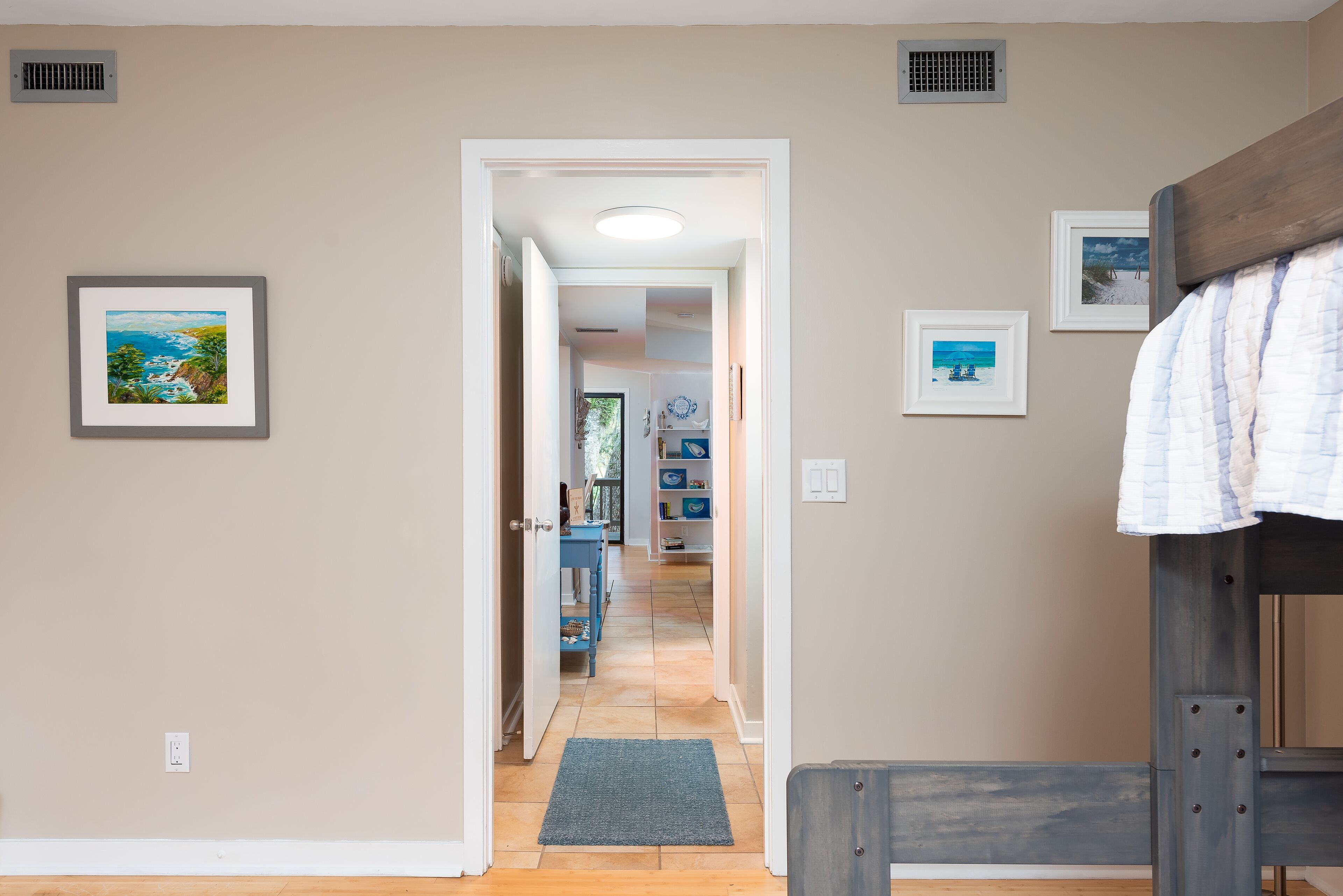 A hallway with a wooden floor, a gray rug, and a bed with a white and blue quilt. There are framed pictures on the walls and a vent on the ceiling.